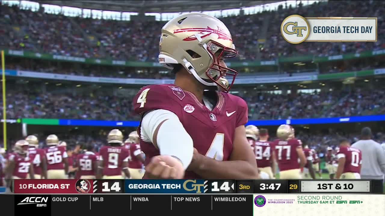 The quarterback, wearing a gold helmet, is looking toward the camera, his arm extended. Behind him, the Florida State Seminoles huddle on the field.
