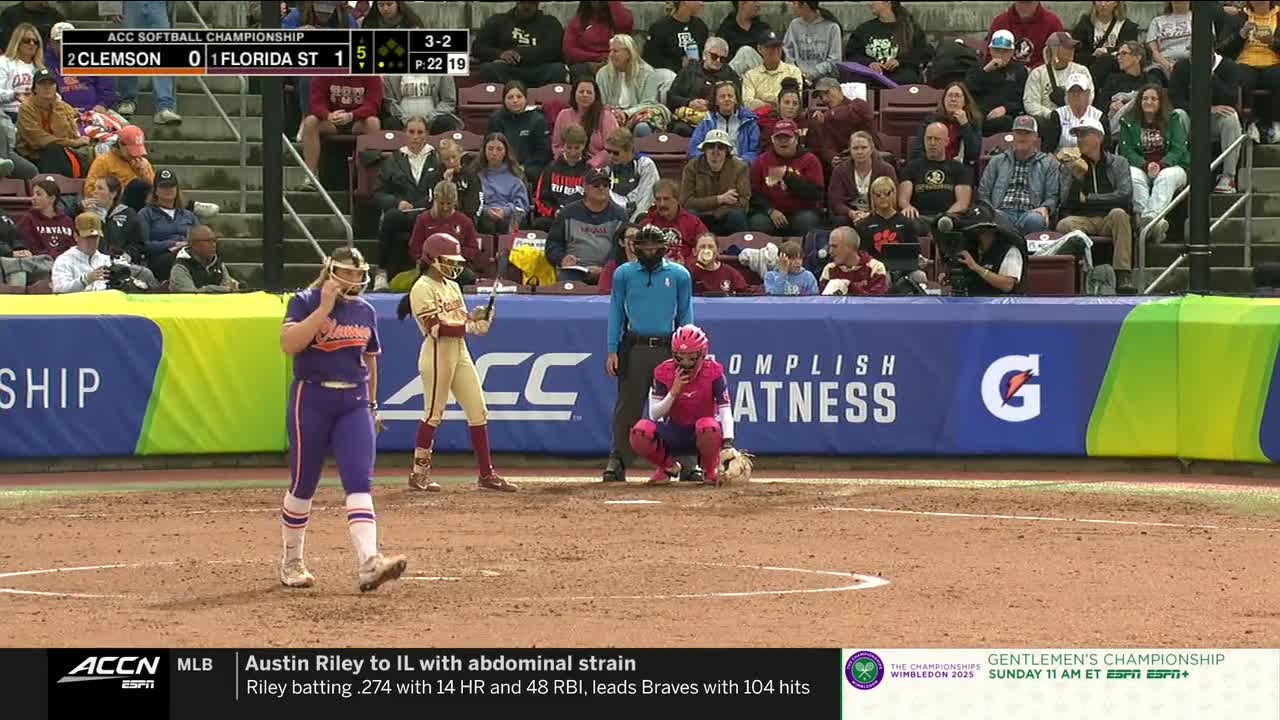 The Clemson pitcher, in purple, walks off the mound, while the Florida State batter stands ready. The umpire and catcher are positioned in front of the plate, awaiting the next play.
