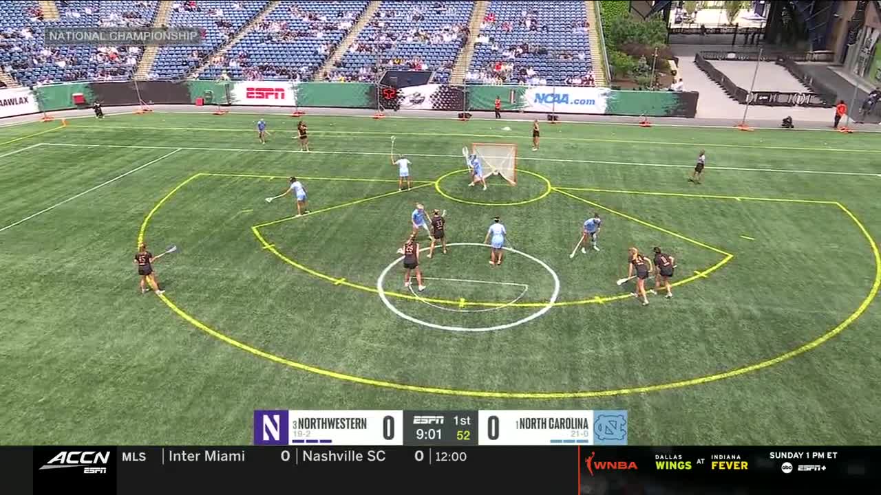 Players in black and light blue uniforms are positioned around the goal, sticks raised. The score is tied, and the clock shows just over nine minutes remaining in the first period.

