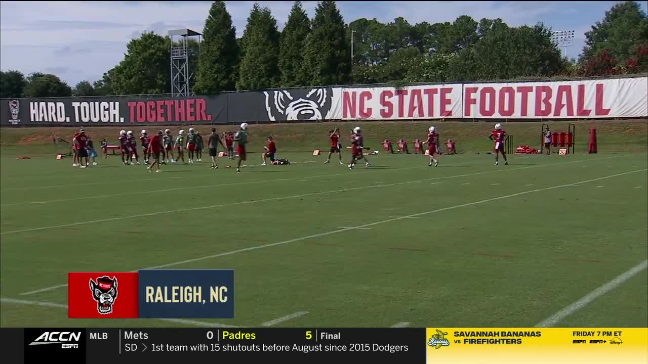 On a bright green field, NC State football players in red and white uniforms are spread out, some stretching and others jogging lightly. Behind them, a banner proclaims "HARD. TOUGH. TOGETHER." and "NC STATE FOOTBALL."
