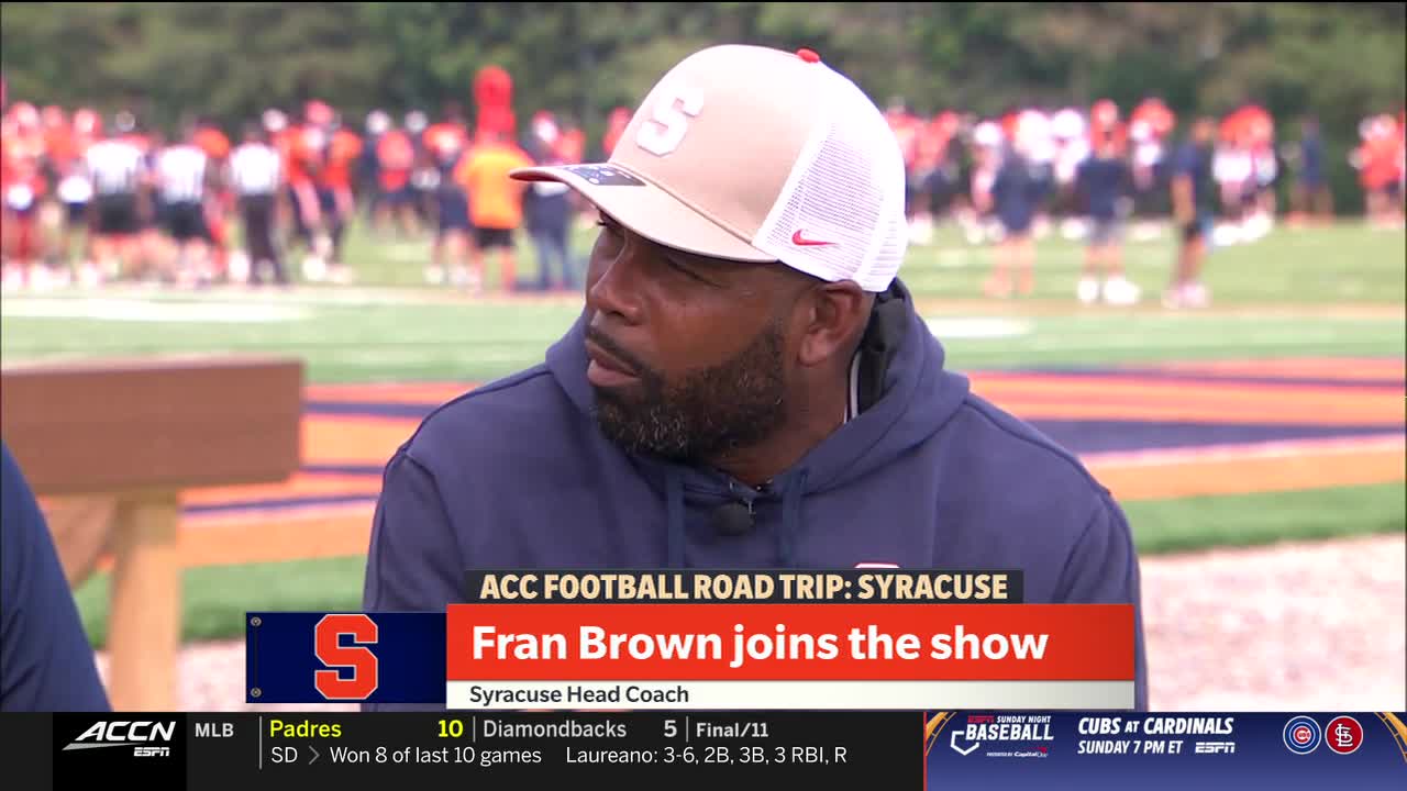 Fran Brown, the Syracuse head coach, is on the ACC Network, wearing an orange and white cap with the Syracuse "S." Beyond him, a blurry group of people in orange and white gear stands on a green field.
