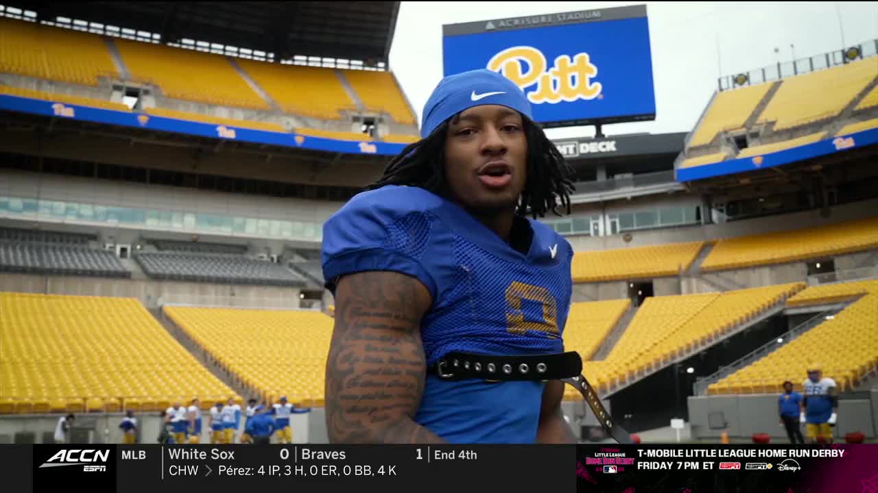 A football player in blue and gold stands on the field, speaking to the camera. Behind him, the stands of Acrisure Stadium are mostly empty, with a few other players visible in the distance.
