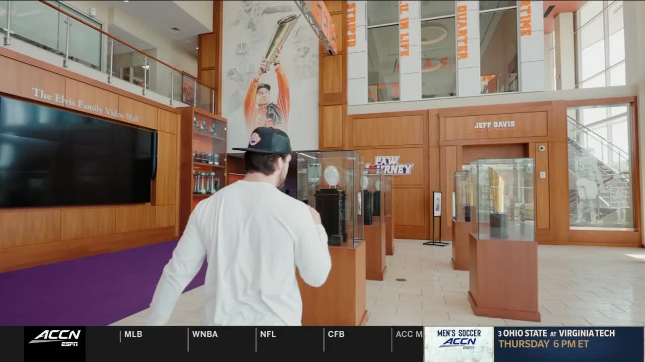 A man wearing a baseball cap walks toward a display of trophies in a wood-paneled room. The ACC Network logo is visible at the bottom of the screen, and a broadcast about Ohio State at Virginia Tech is advertised.
