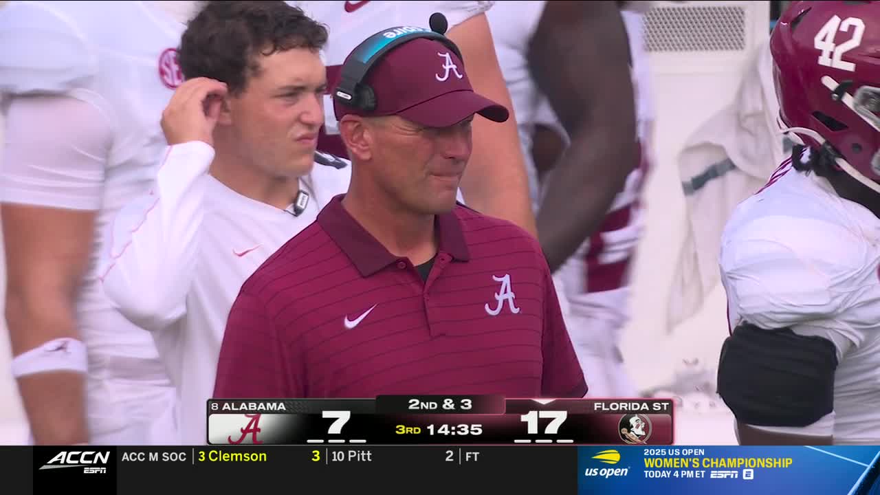 The Alabama coach, wearing a crimson polo and cap, stands on the sidelines, looking down. A player in a maroon helmet and jersey is visible to his right, while another player adjusts his ear to his left.
