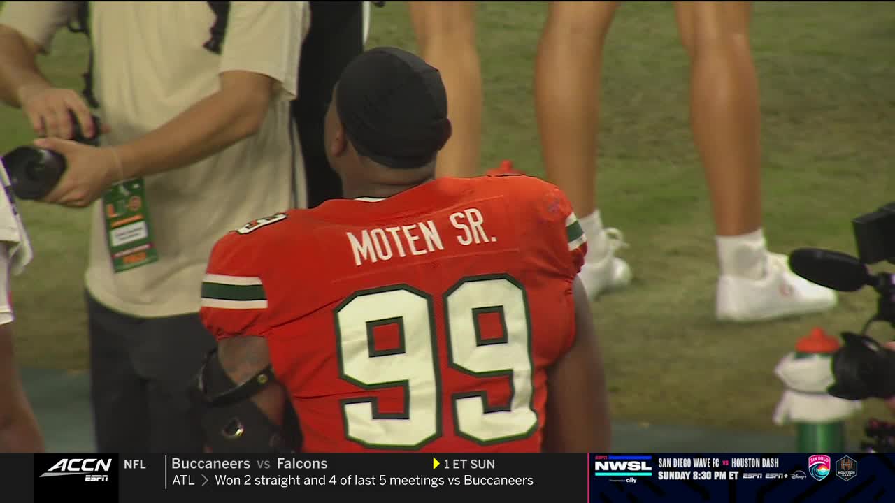 A football player in an orange jersey, number 99, stands facing away, a black cap on his head. A photographer with a camera is positioned to the player's left, while other people stand nearby.
