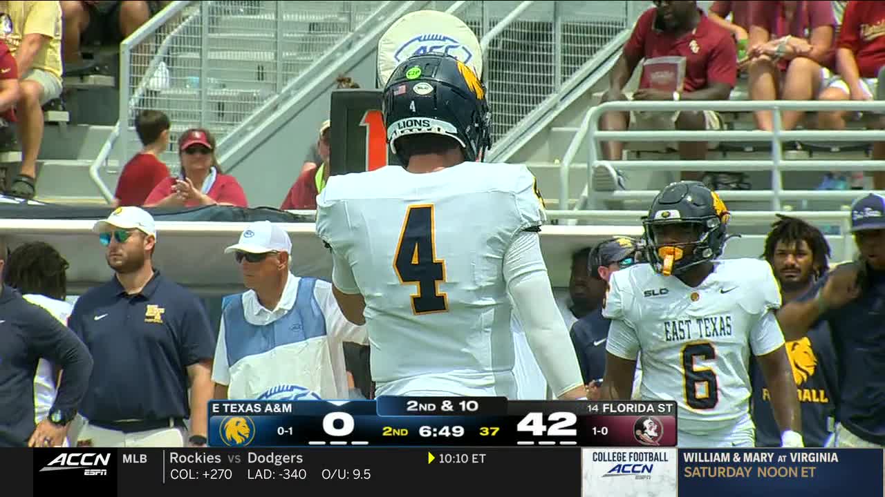 The quarterback, number four, stands with his back to the camera. Across the field, the East Texas player in the number six jersey looks on.
