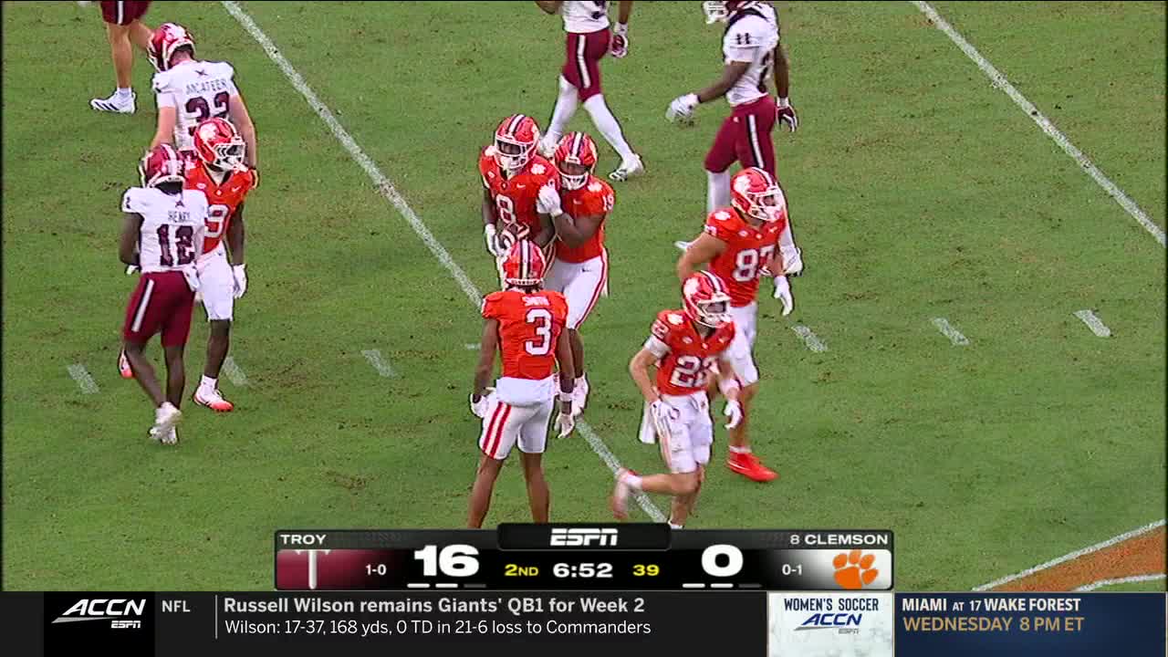 The Clemson players in orange are huddled together, celebrating on the field. A Troy player in maroon stands nearby, watching the action.
