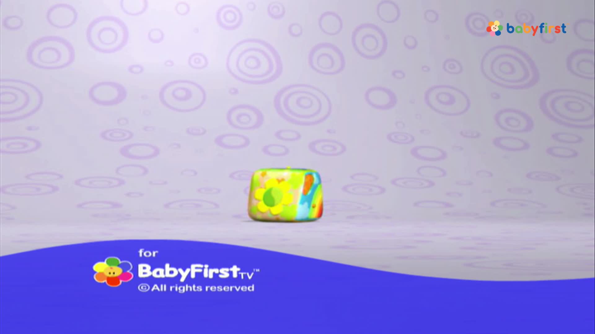 A colorful, patterned cube sits centered on a pale purple background. The BabyFirst logo is at the bottom of the screen.
