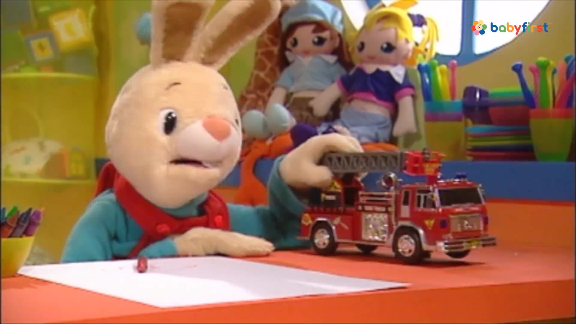 A plush bunny holds a red toy fire truck, its ladder extended. A crayon sits on a blank white paper in front of the bunny.
