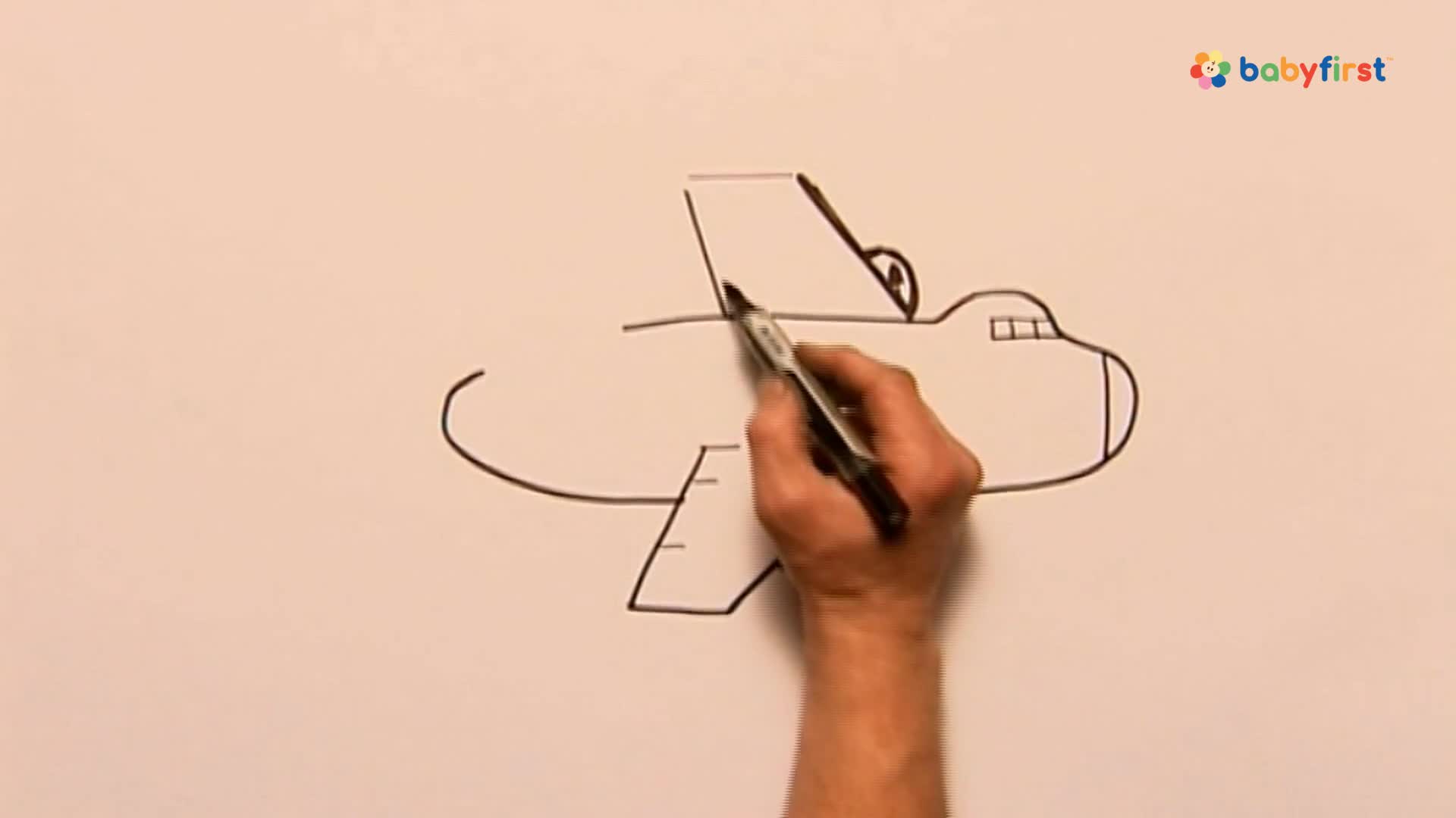A hand holds a black marker, carefully adding lines to a simple airplane drawing. The plane's tail fin and the top of the fuselage are being drawn.
