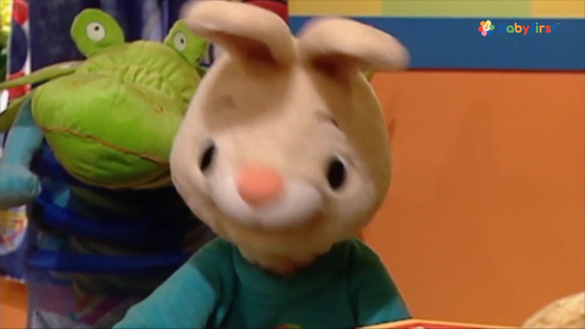 A tan bunny puppet with big black eyes is looking down and to the side. Beside it, a green frog puppet is partially visible.
