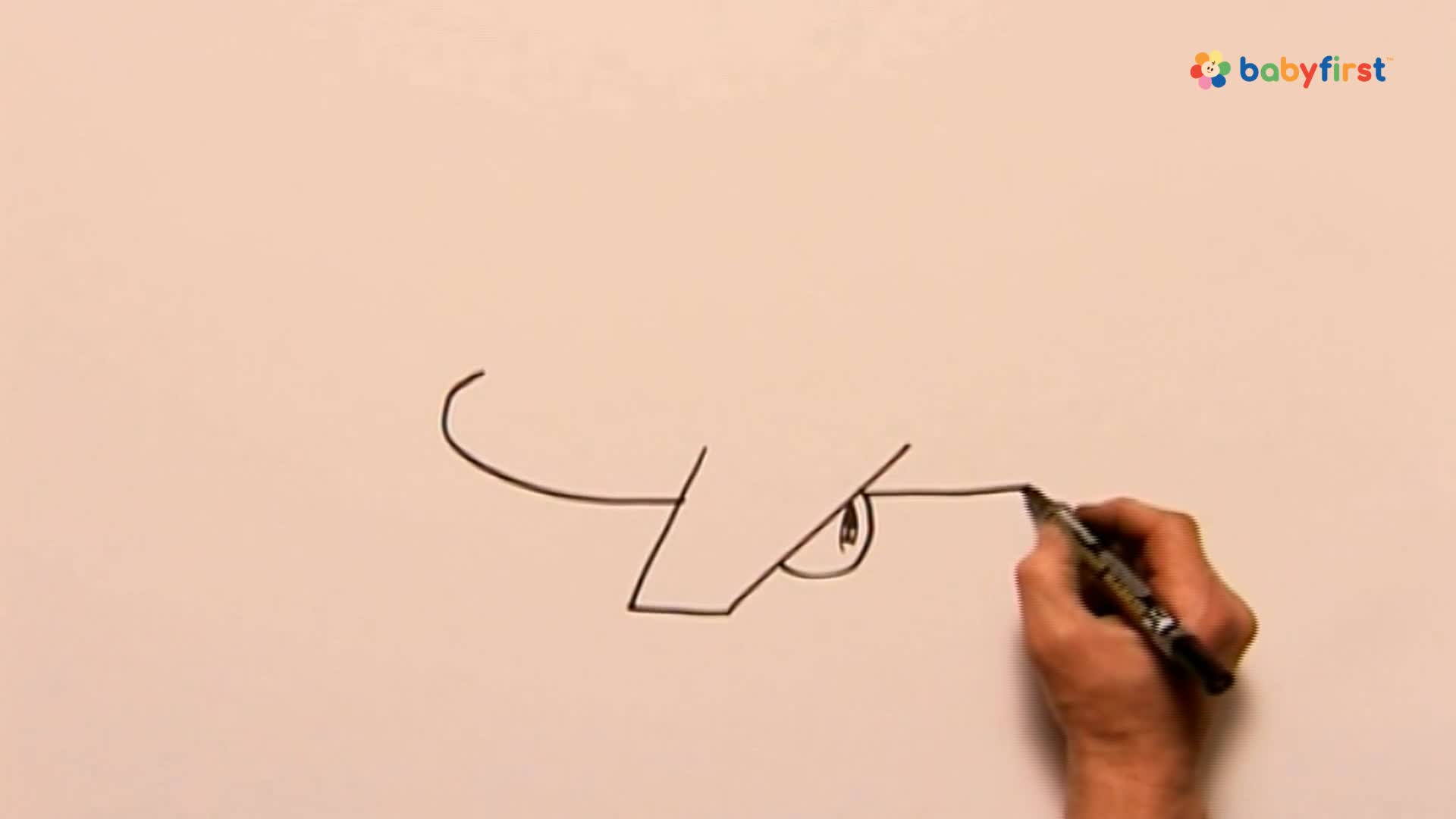 A hand with a black marker is extending the line of a cartoon face. The drawing is being created on a white surface, probably for a BabyFirst show.

