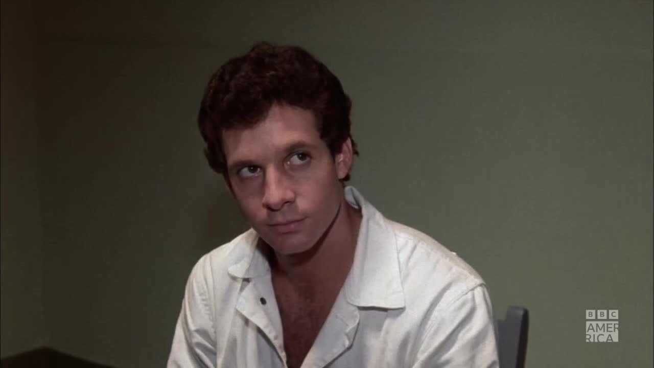 A man sits in a stark room, wearing a white shirt that's unbuttoned at the collar. His gaze is fixed upward, as if considering something unseen.

