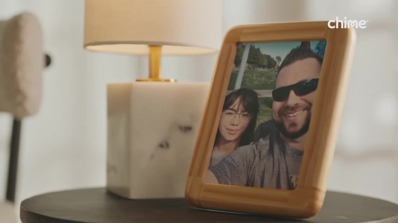 A framed photo of a smiling couple rests on a dark table, next to a glowing lamp. The light catches the gold base of the lamp, and the Chime logo is visible in the corner.
