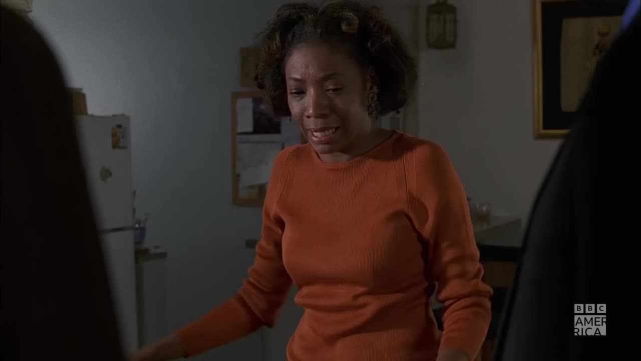 A woman in an orange sweater gestures with her hands, her face etched with concern. The scene unfolds in a kitchen, with a refrigerator and framed artwork visible in the background.
