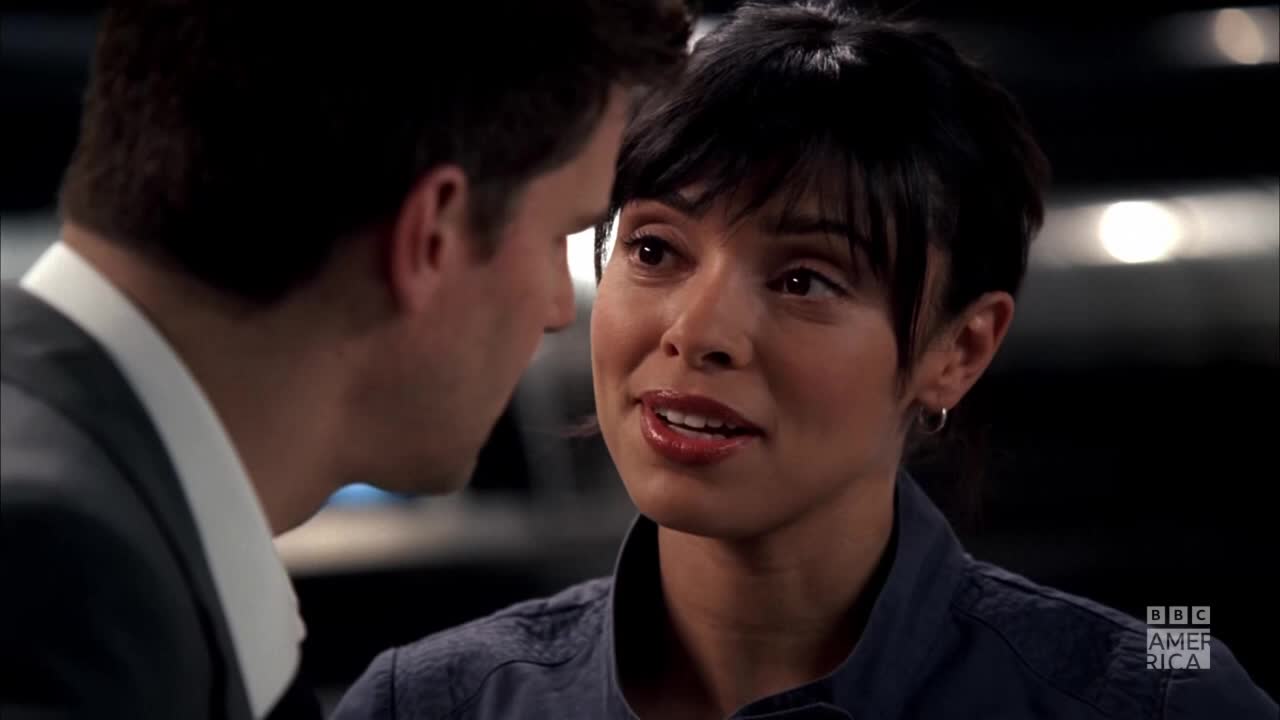 A woman with dark hair and a blue shirt is speaking, her lips slightly parted. Across from her, a man in a suit listens intently, his face partially obscured.
