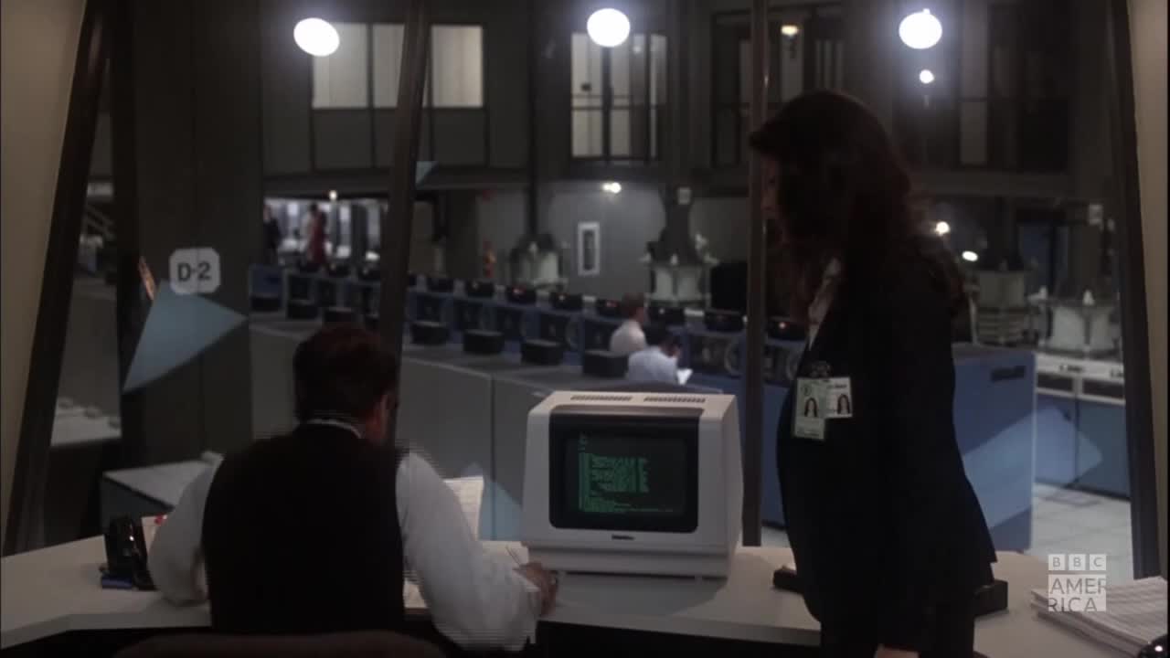 A man hunches over a computer, the screen displaying green text, while a woman stands beside him. Beyond the glass, rows of other computer terminals and people are visible in a large, dimly lit room.
