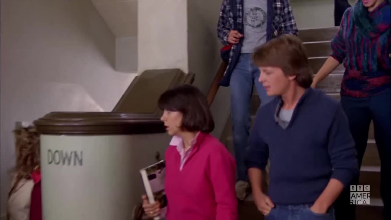 A woman in a pink sweater looks over her shoulder as she descends the stairs, clutching a book. A young man in a blue sweater walks beside her, hands in his pockets, while others follow close behind.
