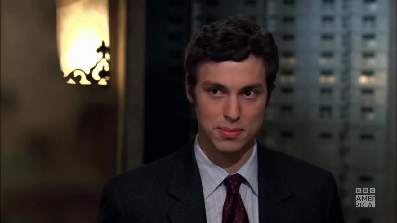 A man in a dark suit looks directly at me, a subtle smirk playing on his lips. The BBC America logo is visible in the corner, suggesting this is a scene from a show.
