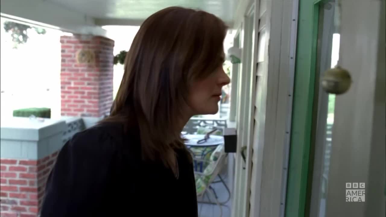 A woman stands near a door, her gaze fixed on something outside. A small, round object hangs near the glass, and the BBC America logo is visible on the right.

