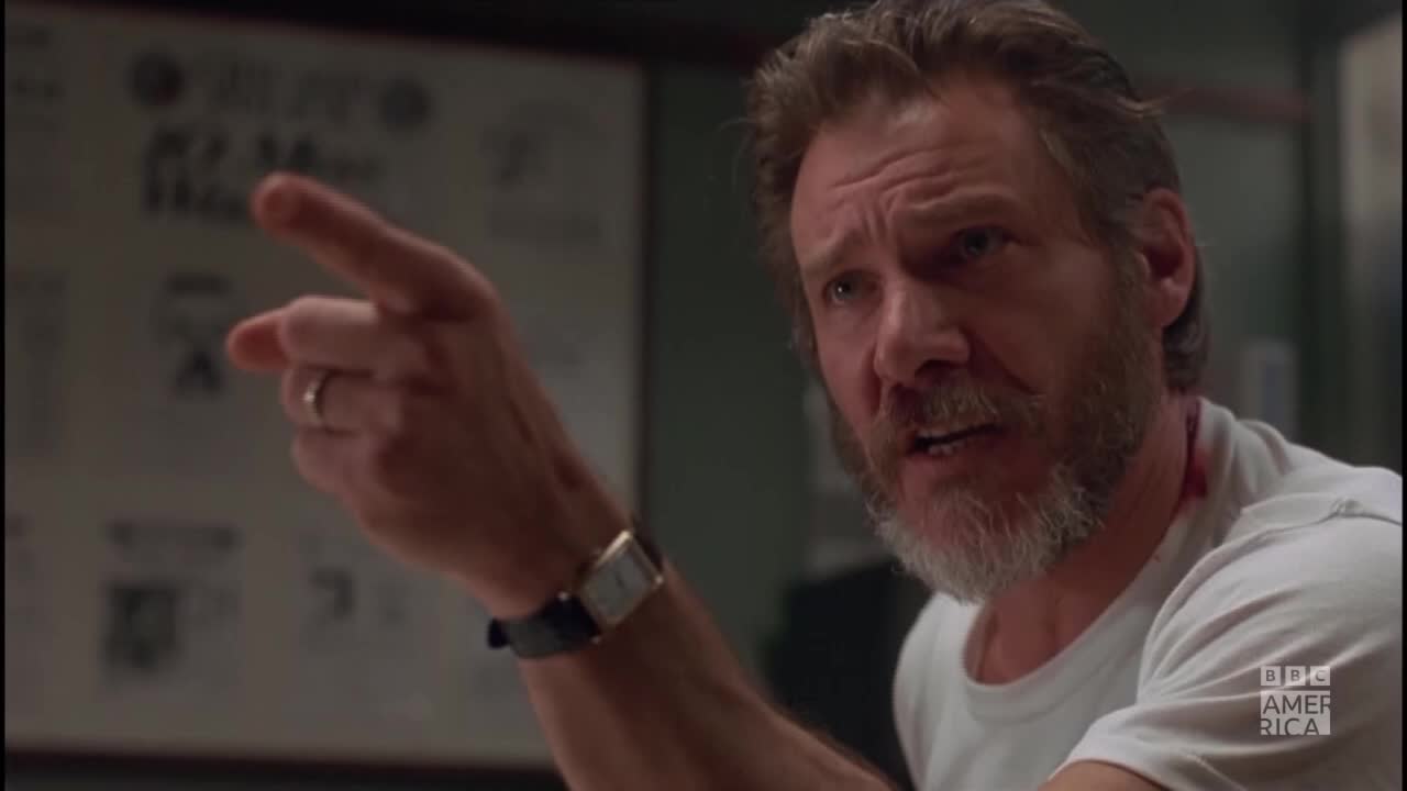 A man with a gray beard and a white t-shirt gestures emphatically with his right hand. His index finger is extended, and he's pointing toward something off-screen.
