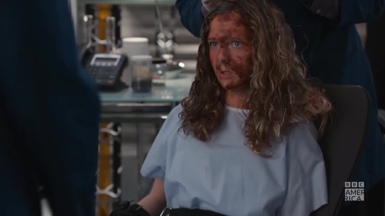 A woman with blood smeared across her face sits in a chair, staring forward. She's wearing a hospital gown, and the BBC America logo is visible in the corner.
