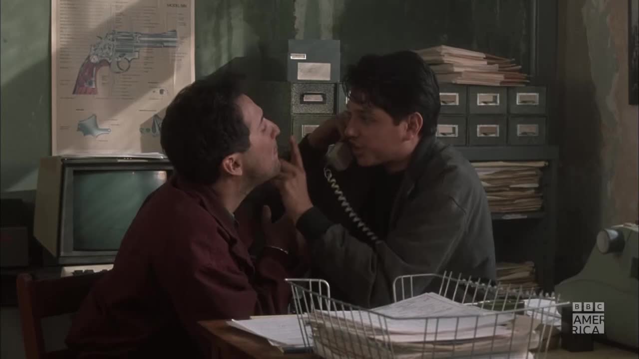 A man in a dark jacket gestures emphatically while speaking on the phone, his finger nearly touching the face of the man beside him. The scene, likely from a BBC America show, unfolds in a cluttered office with a diagram of a gun on the wall.
