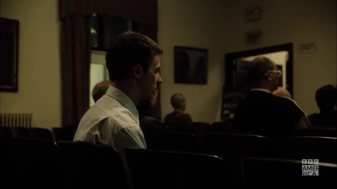 A young man in a white shirt sits in a dark room, his gaze fixed forward. Other people are seated around him, and the BBC America logo is visible in the corner.
