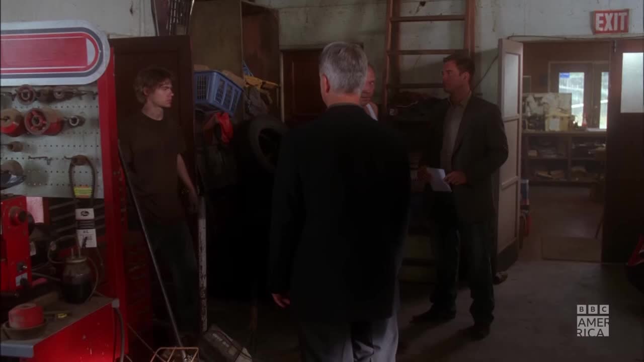 A man in a dark suit faces away from me, talking to two other men in a cluttered garage. A young person in a t-shirt stands in a doorway, watching the conversation.
