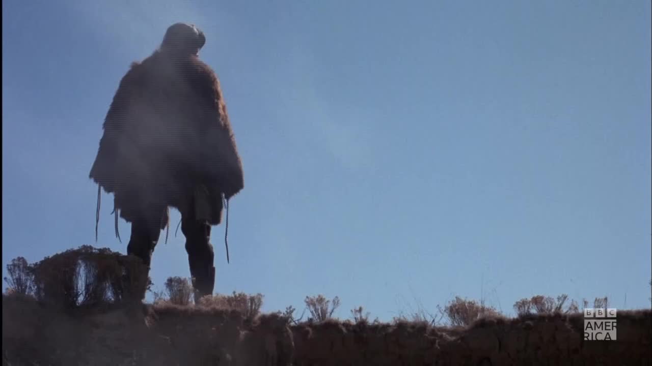 A figure, cloaked in brown fur, stands silhouetted against a bright blue sky. The BBC America logo is visible in the corner.
