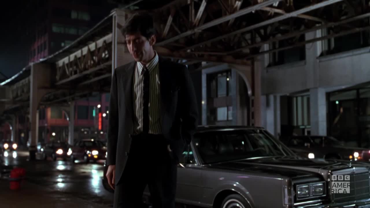 A man in a suit stands next to a classic car on a city street at night. Headlights from passing cars reflect off the wet pavement under the elevated train tracks.
