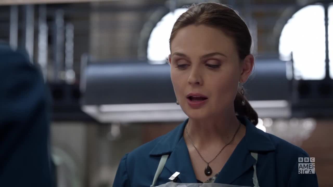A woman in a blue lab coat is speaking, her mouth open as if mid-sentence. Behind her, a blurry industrial setting suggests a scene from the United States, possibly on BBC America.
