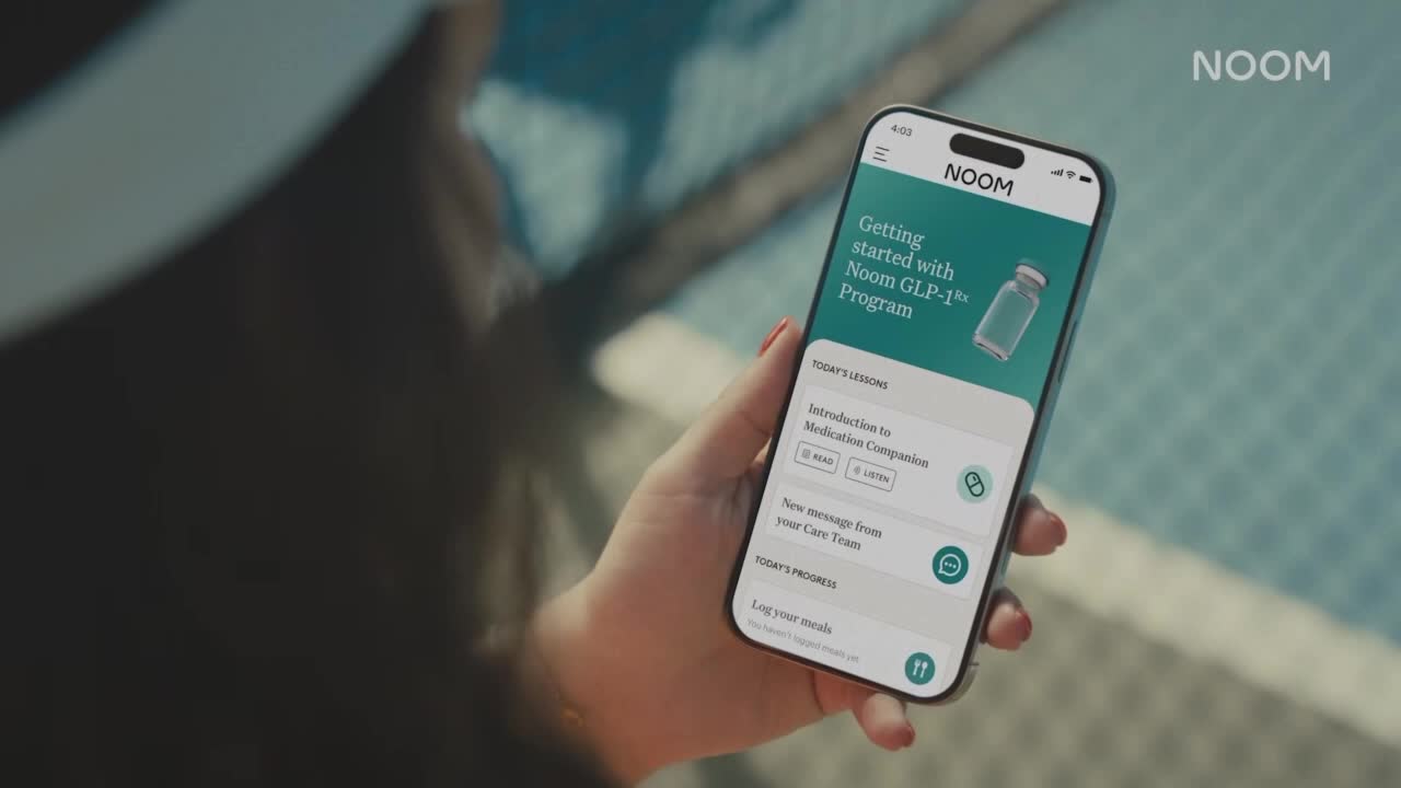 A hand holds a phone displaying the Noom app, its screen reflecting the sunlight. The app's interface shows options for a GLP-1 program.
