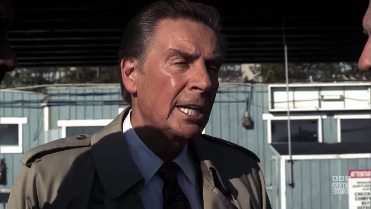 The man in the trench coat is speaking, his mouth open as he gestures with his hands. The BBC America logo is visible in the background.
