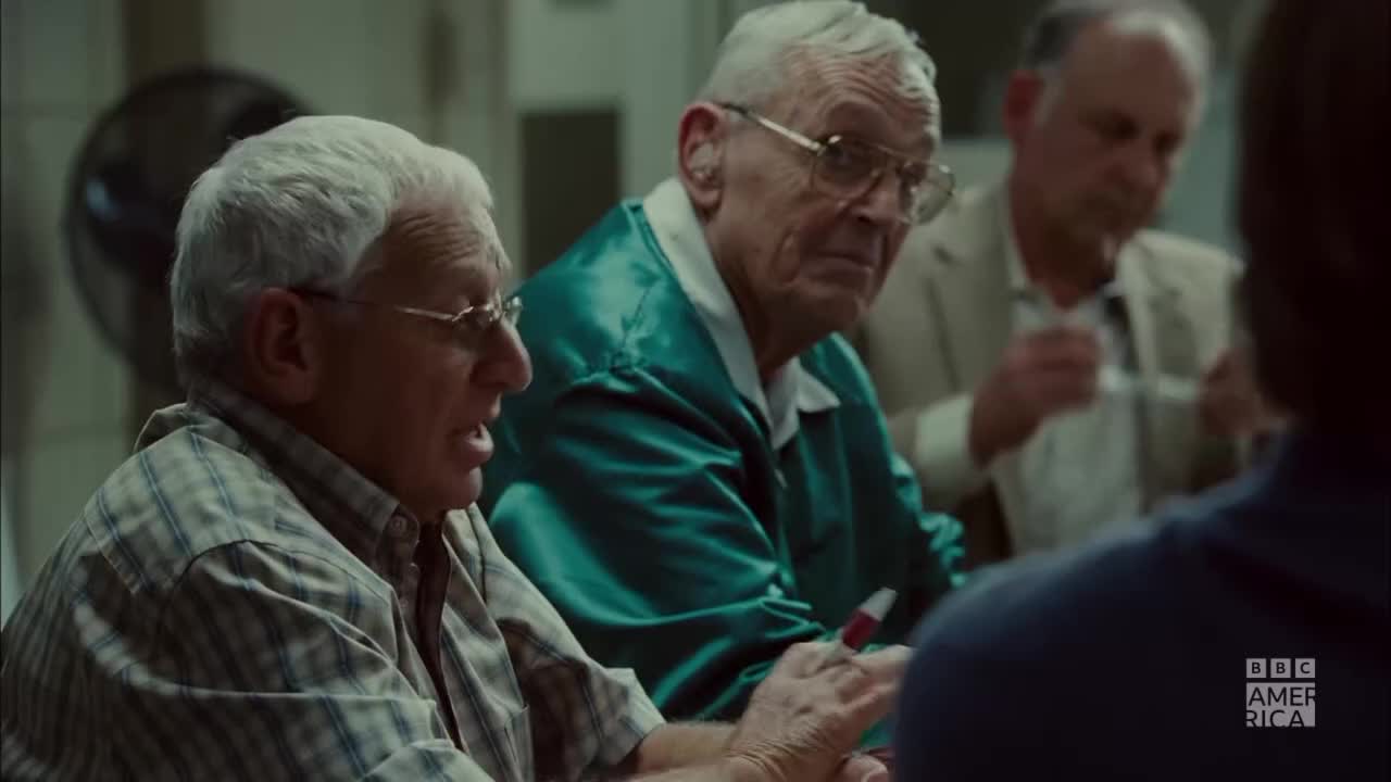 A man in a plaid shirt gestures emphatically, his mouth open as he speaks. Beside him, another man in a green jacket looks on, a red marker held loosely in his hand.
