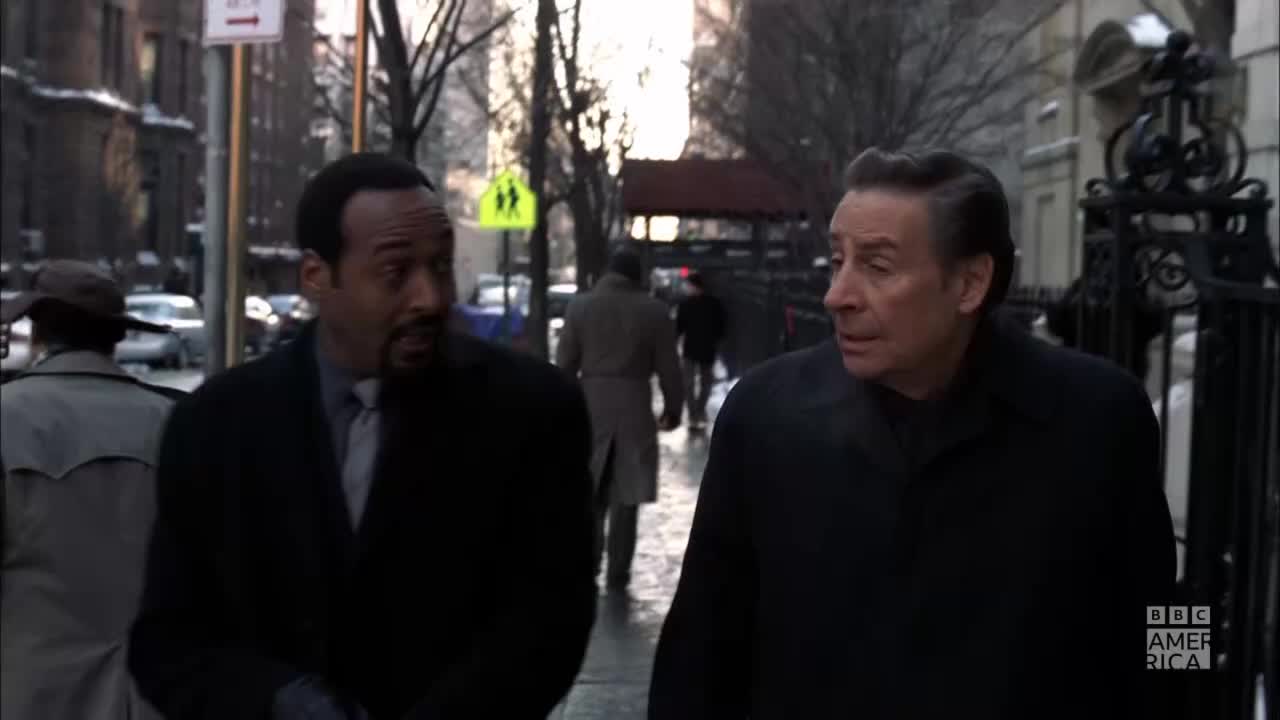 Two men in dark coats walk side-by-side on a city sidewalk. The man on the left, wearing a tie, appears to be speaking to his companion.
