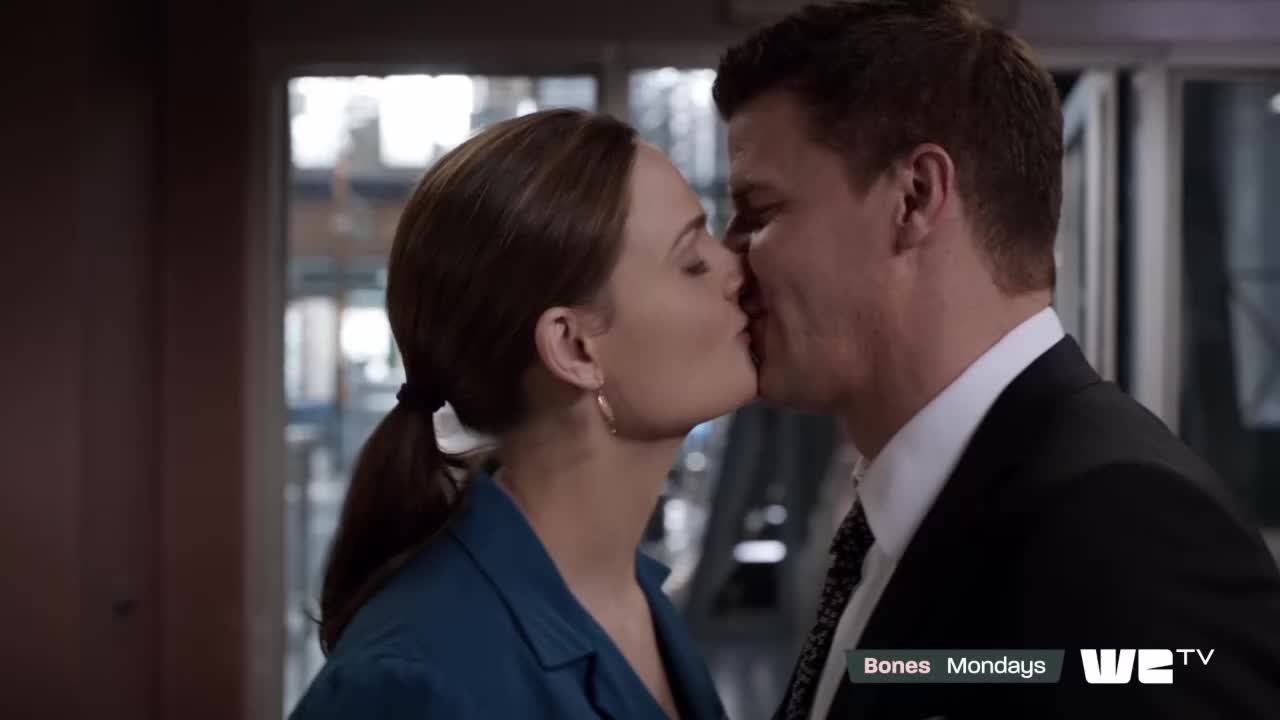 A man in a suit and a woman in a blue jacket are locked in a kiss. The woman's hair is pulled back in a ponytail.
