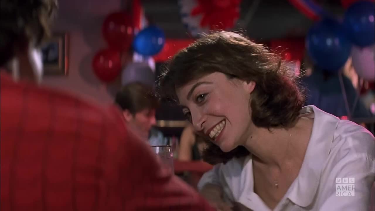 A woman in a white blouse is smiling, leaning towards someone out of frame. Red, white, and blue balloons hang in the background, and the BBC America logo is visible.
