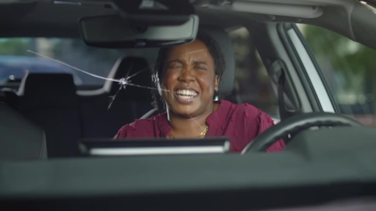 The woman in the driver's seat is laughing, her face lit up, despite the cracked windshield. The spiderweb of damage spreads from a point above her head.

