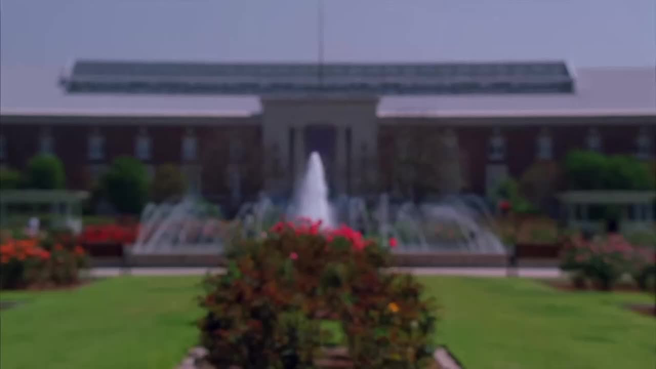 Water jets shoot high into the air from a fountain in front of a large building. A rose garden frames the foreground, and the scene is bathed in soft light.
