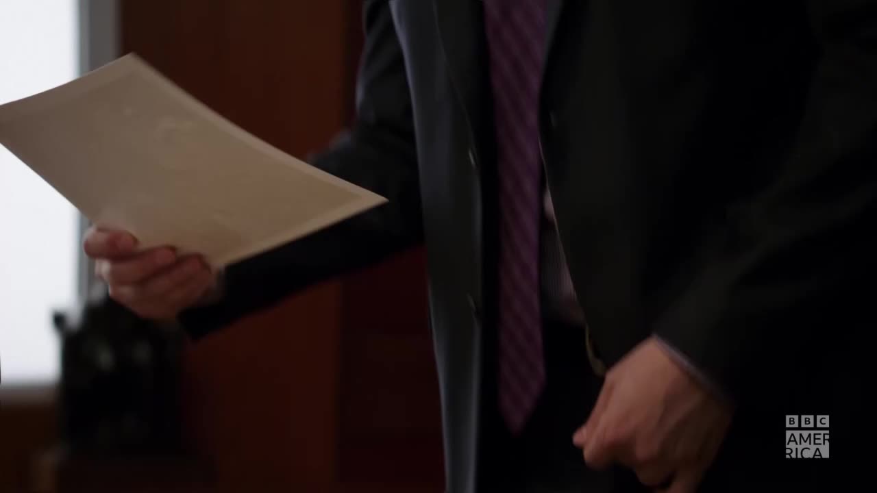 A man in a dark suit holds a large, cream-colored envelope. His purple-striped tie is visible as he reaches toward something out of frame, perhaps to hand it off.
