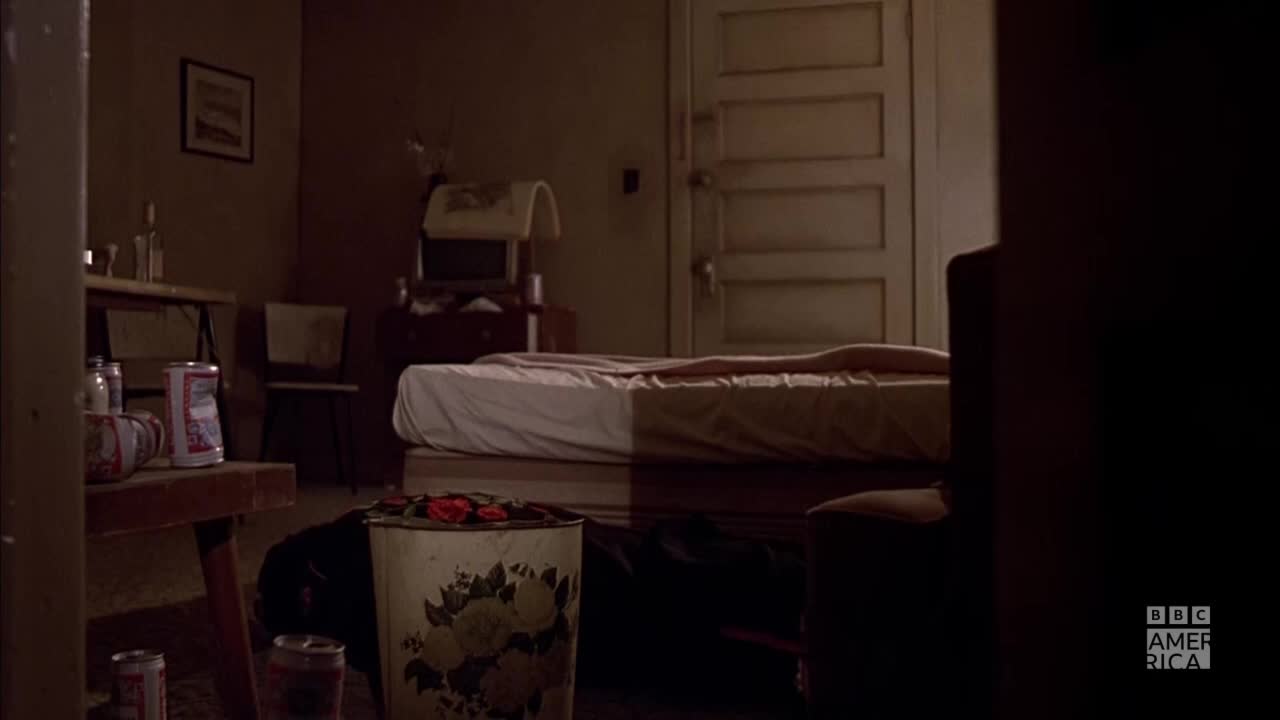 A bed is illuminated by a single light source, with a figure lying beneath the covers. The room is dimly lit, with discarded beer cans and a floral waste bin visible in the foreground.
