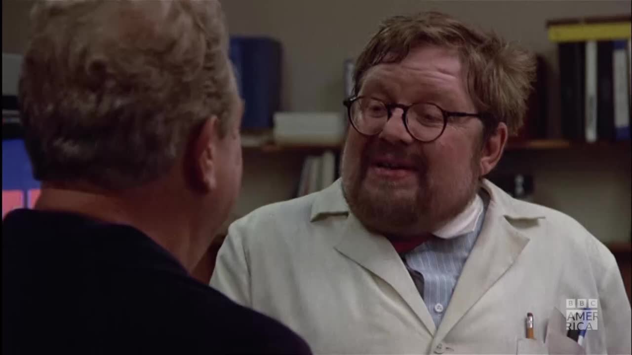 A man in a lab coat and round glasses is speaking intently, his face animated. The other man, in a dark shirt, is listening, his back to the camera.
