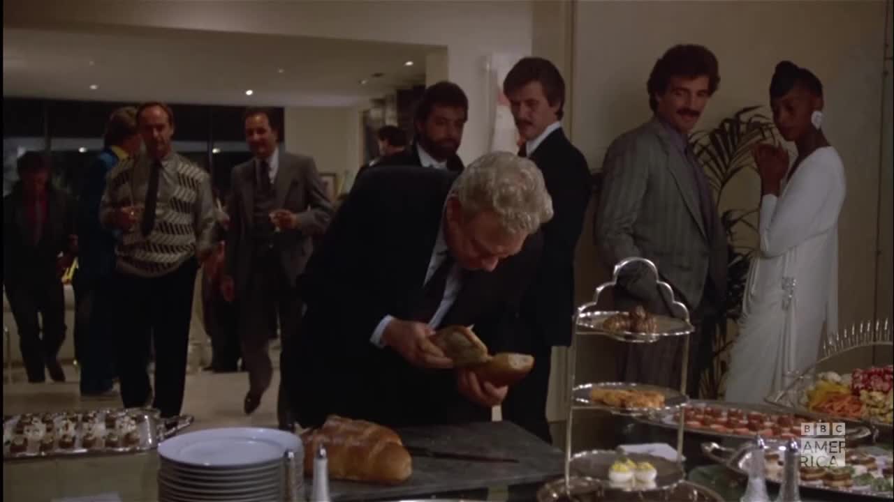 A man in a dark suit carefully cuts into a loaf of bread on a buffet table. Guests in formal attire mingle nearby, some holding drinks.

