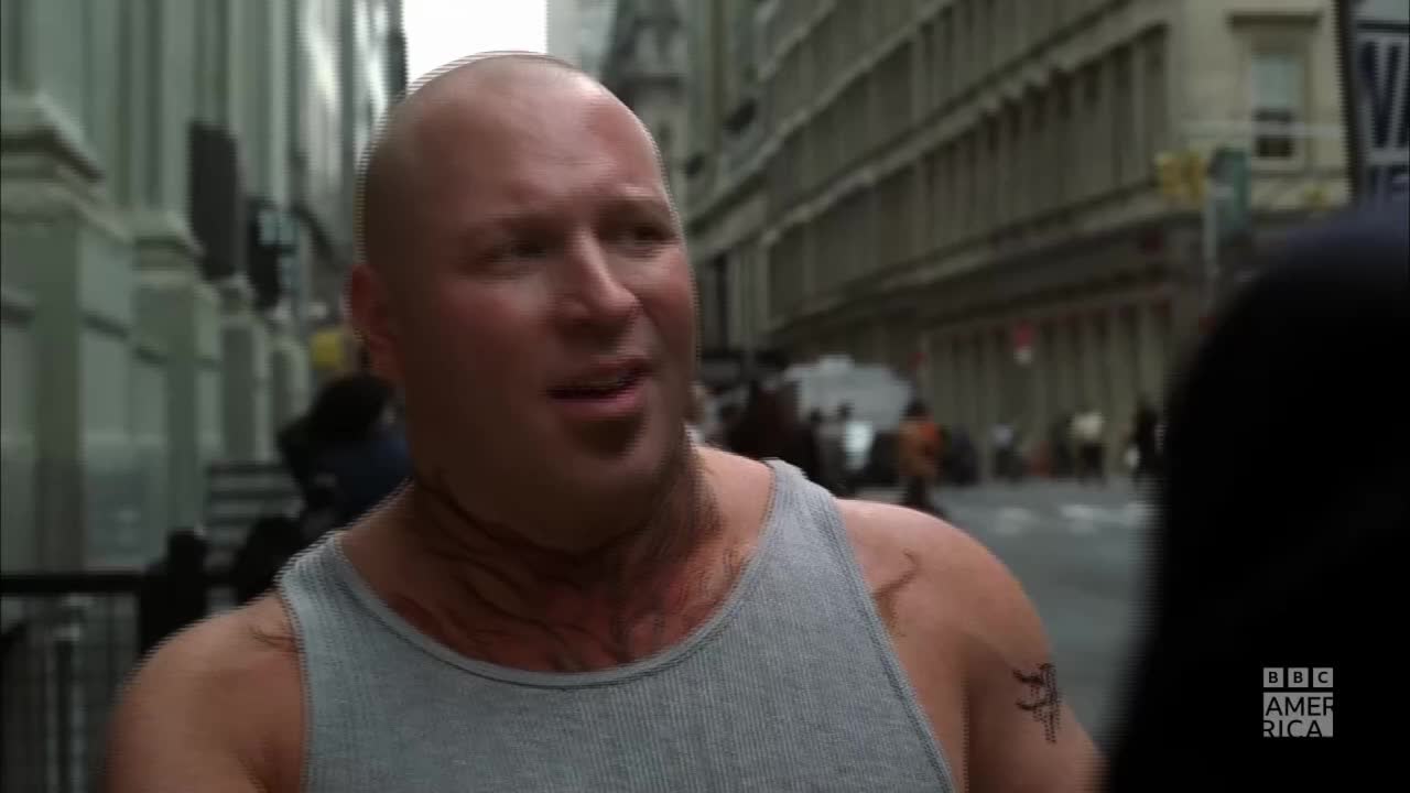 A bald man in a gray tank top is speaking, his face animated with emotion. He’s standing on a city street, with buildings and people in the background, a BBC America logo in the corner.
