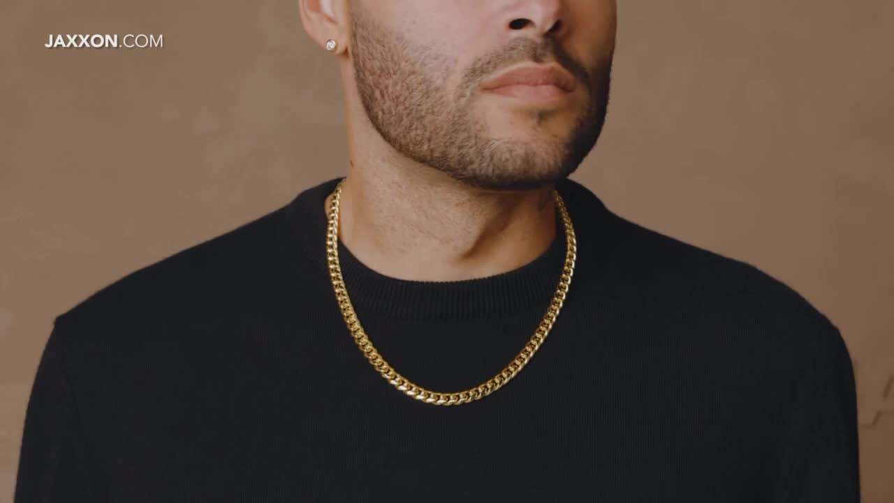 A man with a diamond stud earring and a gold chain necklace is centered in the frame. He wears a black sweater against a neutral background.
