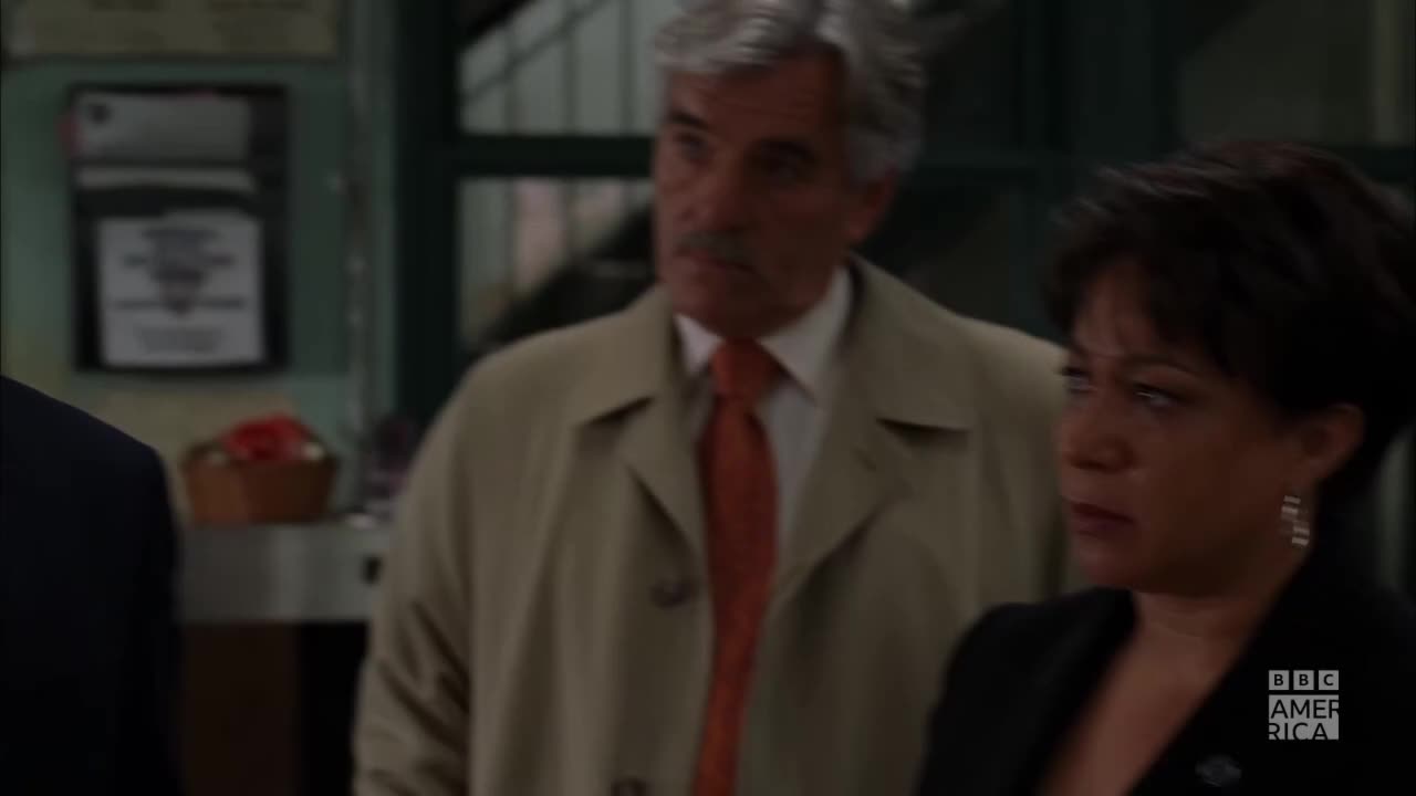 A man in a tan coat and orange tie looks off to the side, while a woman in a black blazer stares forward. The BBC America logo is visible on the right.
