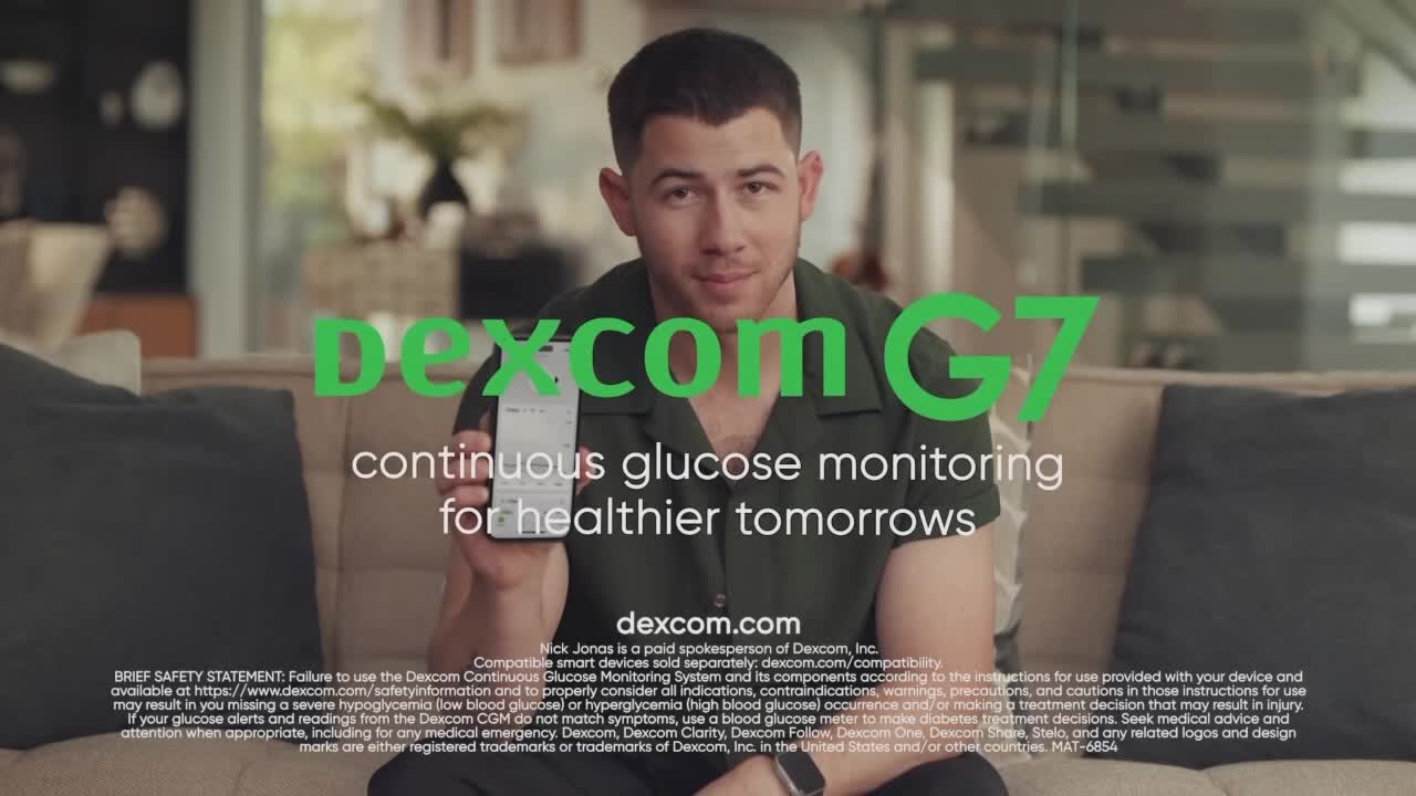Nick Jonas sits on a couch, holding a phone displaying a glucose monitoring app. The Dexcom G7 logo and tagline are prominently displayed over him.

