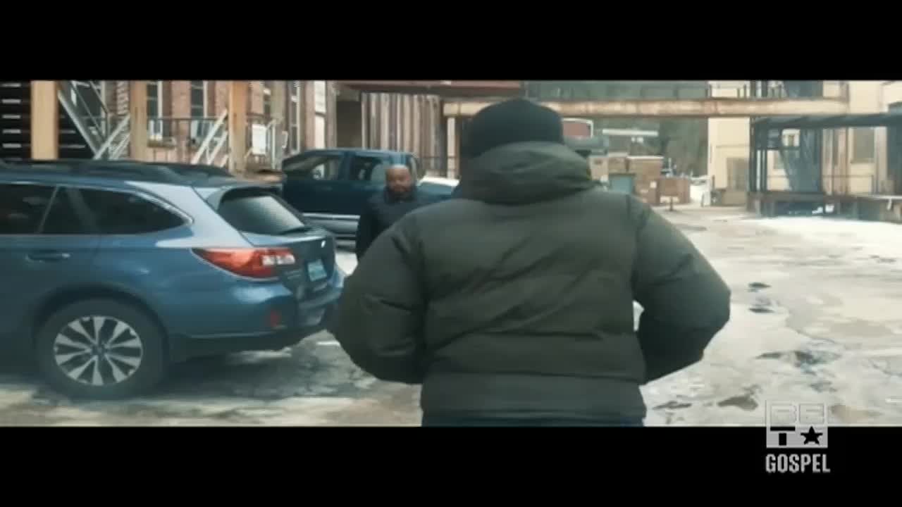 A man in a puffy green coat walks away from the camera, heading toward a blue SUV. Another man in a black coat is walking in the same direction, further down the snowy path.
