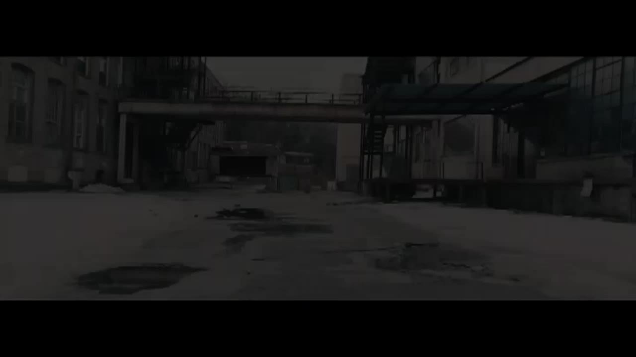 The camera is positioned in a bleak, industrial courtyard, the ground wet and reflecting the dim light. A metal bridge connects two buildings, casting a shadow over the space.
